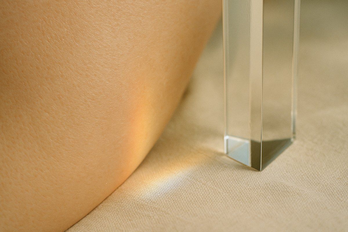Macro still life of a glass prism on ivory linen beside a fragment of real skin, warm daylight refracting into a soft caustic, broadband light editorial at Facial Sculpting London