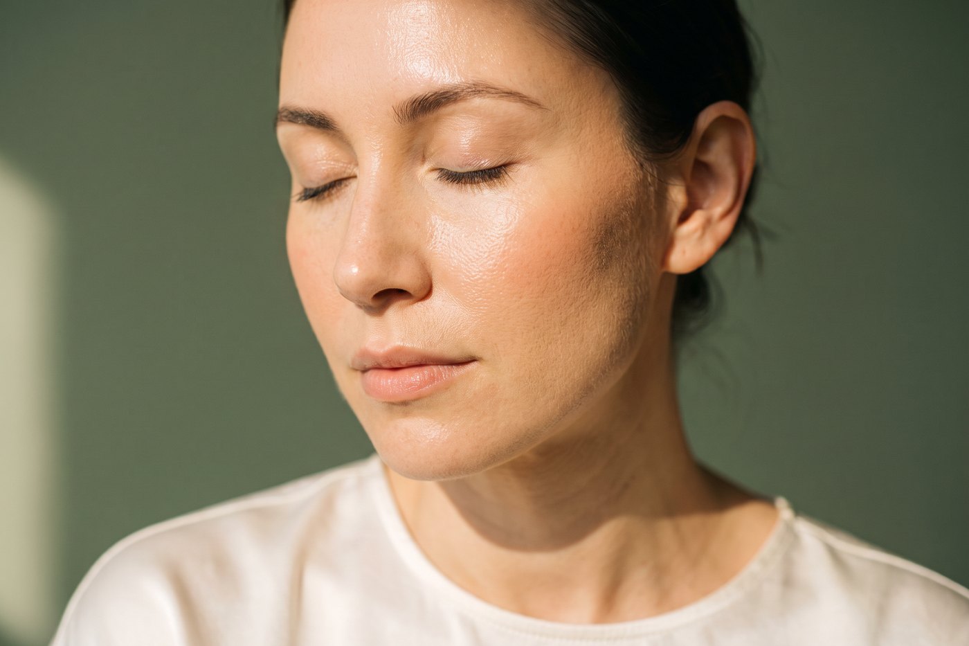 Serene beauty editorial portrait of a woman with luminous glass-skin glow in soft warm light against sage green, the skin quality SkinPen microneedling restores at Facial Sculpting London
