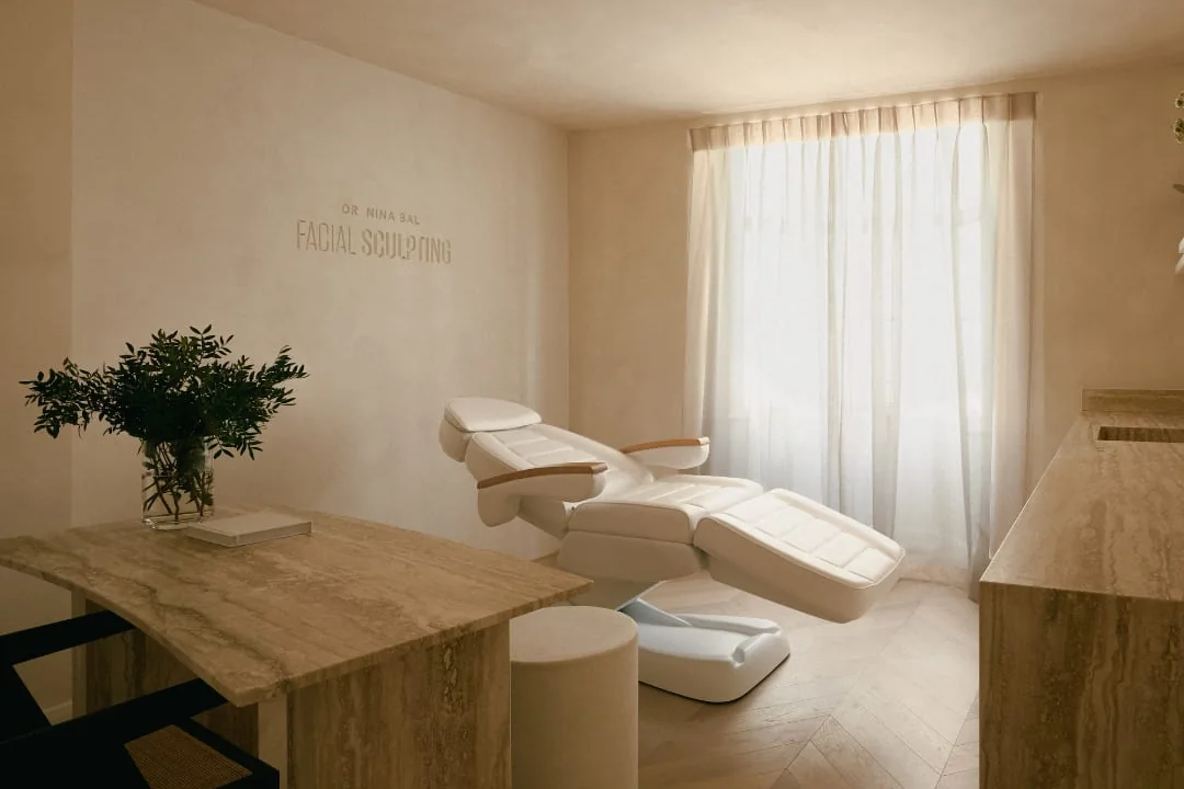 A treatment room at the Facial Sculpting clinic in South Kensington, with the treatment chair by a bright curtained window and the clinic name on the wall